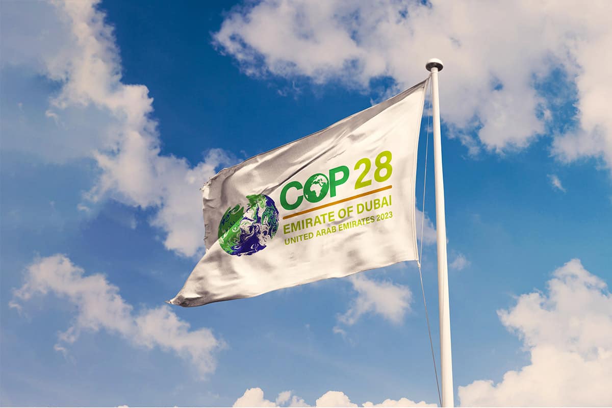 Fahne Klimakonferenz COP28(c)Adobestock/Rafael Henrique Eine weiße Fahne weht im Wind vor blauem Himmel und wenigen weißen Wolken. Auf der Fahne steht COP28, Emirate of Dubai. United Arab Emirates 2023. Links neben der Schrift ist eine stilisierte Erdkugel in grün-blau zu sehen.