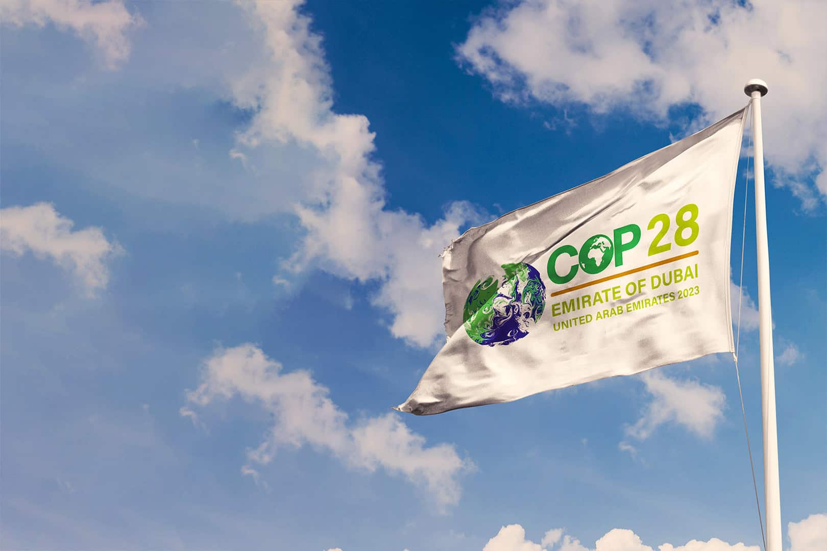 Fahne Klimakonferenz COP28(c)Adobestock/Rafael Henrique Eine weiße Fahne weht im Wind vor blauem Himmel und wenigen weißen Wolken. Auf der Fahne steht COP28, Emirate of Dubai. United Arab Emirates 2023. Links neben der Schrift ist eine stilisierte Erdkugel in grün-blau zu sehen.